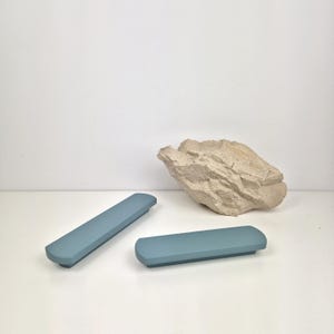 Puede incluir: Dos tiradores de armario rectangulares azul claro con extremos redondeados sobre una superficie blanca. Una roca beige texturizada está en el fondo. Los tiradores miden unos 20 cm de largo.
