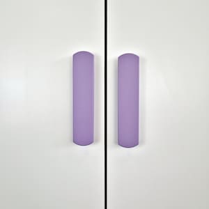 Könnte beinhalten: Nahaufnahme einer weißen Schranktür mit zwei vertikalen, lavendelfarbenen Schrankgriffen. Die Griffe sind oben und unten abgerundet und gleichmäßig beabstandet.