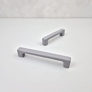 May include: Two gray metal cabinet pulls with a rectangular shape and rounded edges. The pulls are shown on a white surface.
