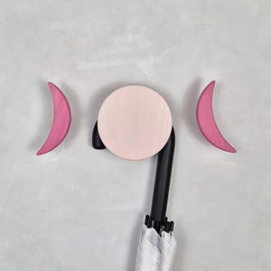 Può includere: Un gancio decorativo da parete con un cerchio rosa chiaro e due forme di luna crescente rosa. Il gancio è attaccato a un manico di ombrello nero, con l'ombrello bianco parzialmente visibile. Il design è su uno sfondo grigio chiaro.