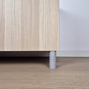 May include: A light-coloured wooden cabinet with a grey base and grey cylindrical legs. The cabinet is set against a white wall and a light brown wooden floor.