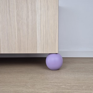 Puede incluir: Un mueble de madera de color claro con una sola pata redonda de color lavanda. El mueble descansa sobre un suelo de madera marrón claro. La pared detrás del mueble es blanca.