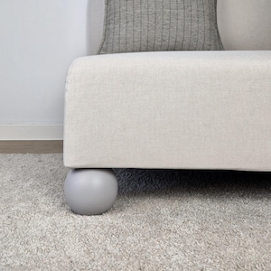 May include: A section of a cream-coloured sofa with a grey, spherical foot. A grey patterned cushion rests on the sofa. The sofa sits on a light grey carpet.