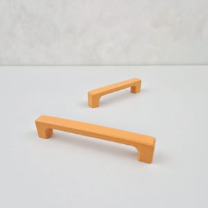 Puede incluir: Dos tiradores de armario naranjas con forma rectangular. Los tiradores están hechos de un material liso y sólido.
