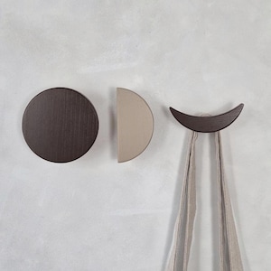 May include: Three wooden wall hooks in a minimalist design. One is a dark brown circle, another is a half-circle in beige, and the third is a crescent shape in dark brown, with a beige fabric bag hanging from it.