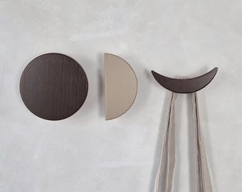 May include: Three wooden wall hooks in a minimalist design. One is a dark brown circle, another is a half-circle in beige, and the third is a crescent shape in dark brown, with a beige fabric bag hanging from it.