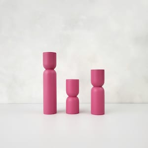 Könnte beinhalten: Drei zylindrische Kerzenhalter in Pink, in verschiedenen Höhen. Die Kerzenhalter sind aus Holz und haben eine glatte, matte Oberfläche.