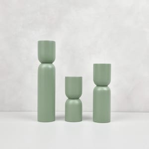 Può includere: Tre portacandele verdi di altezze diverse. I portacandele sono in legno e hanno una forma cilindrica con un leggero design a clessidra.