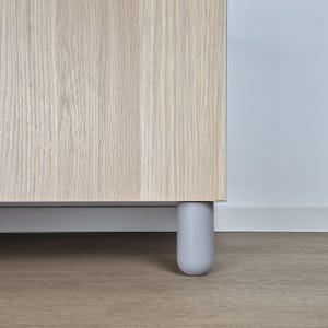May include: Close-up of a light-coloured wooden cabinet with a grey, rounded leg. The cabinet has a natural wood grain texture. The leg is cylindrical with a rounded bottom. The cabinet is set against a light-coloured wall and floor.