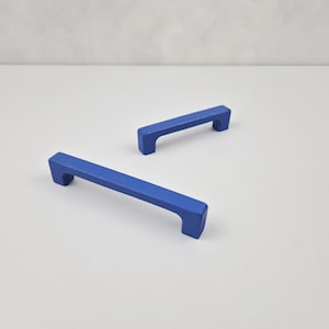 Könnte beinhalten: Zwei blaue Schrankgriffe mit rechteckiger Form und abgerundeten Kanten. Die Griffe sind aus Holz und haben eine matte Oberfläche.