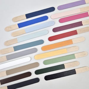 May include: A collection of wooden paint stirrers, each with a different color painted on one end. Colors include navy, blue, light blue, teal, gray, white, tan, brown, black, purple, pink, magenta, burgundy, red, orange, yellow, green, and dark green.