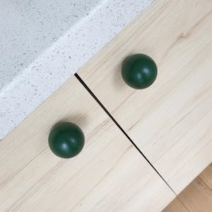 May include: Two dark green spherical cabinet knobs on a light wood cabinet door. The knobs are evenly spaced and positioned diagonally. The cabinet door has a natural wood grain. A white speckled countertop is visible in the background.