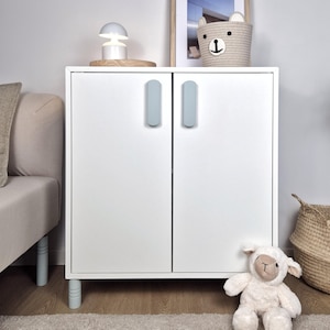 May include: A white cabinet with two doors and light blue handles. The cabinet has light blue legs and is next to a beige sofa. A stuffed lamb and a woven basket are on the floor. A lamp and a bear-shaped basket sit on top of the cabinet.