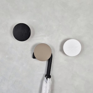 May include: Three round wall hooks in black, beige, and white. The black hook has a wood grain texture. A black and white umbrella hangs from the beige hook. The hooks are mounted on a light gray wall.