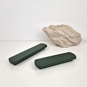May include: Two dark green rectangular cabinet pulls with rounded edges are displayed on a white surface. A large, textured, light-colored rock sits in the background. The pulls are evenly spaced and appear to be made of a solid material.