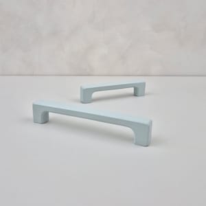 May include: Two light blue cabinet pulls with a rectangular shape. The pulls are made of metal and have a smooth finish.