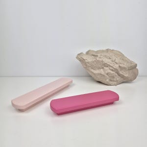 Może przedstawiać: Dwa prostokątne obiekty w pastelowych kolorach, jeden jasnoróżowy, a drugi w żywym kolorze magenta, są prezentowane na białej powierzchni. Teksturowana, beżowa formacja skalna znajduje się w tle, dodając naturalnego kontrastu.