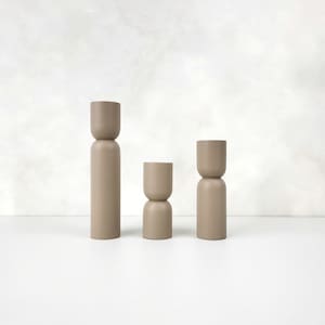 May include: Three beige wooden decorative objects of varying heights. Each object has a cylindrical base and a rounded top, creating a minimalist, modern aesthetic. The objects are arranged on a white surface against a neutral background.