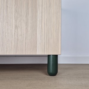 May include: Close-up of a light wood cabinet with dark green cylindrical legs. The cabinet's wood grain is visible, and the legs have a rounded bottom. The cabinet is set on a light wood floor.