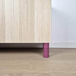 Könnte beinhalten: Ein Teil eines hellfarbenen Holzschranks mit einem einzelnen, zylindrischen, magenta-farbenen Bein. Das Bein hat drei horizontale Rillen. Der Schrank steht vor einer weißen Wand und einem hellen Holzboden.