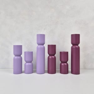 May include: Six wooden decorative objects in varying shades of purple and burgundy. The objects have a unique hourglass and cylindrical design. They are arranged in a row against a neutral background, showcasing their different heights and colors.