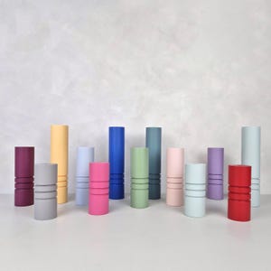May include: Assortment of 14 colorful cylindrical candle holders in various shades of blue, green, pink, red, yellow, and gray. The holders have a grooved design and are arranged in a row on a white surface.