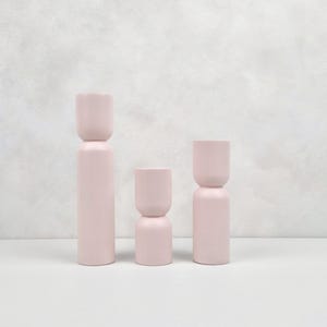 May include: Three pink ceramic candle holders of varying heights. The holders are cylindrical with a slight inward curve at the top and bottom.