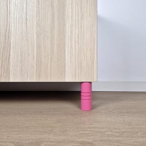 Può includere: Un mobile in legno chiaro con una gamba cilindrica rosa. La gamba ha tre scanalature orizzontali. Il mobile è appoggiato a una parete bianca e a un pavimento in legno marrone chiaro.