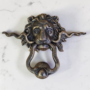 Puede incluir: Aldaba de puerta de color bronce con la cabeza de un león y un asa en forma de serpiente. El león tiene una melena y rasgos faciales detallados. La aldaba es de metal y tiene un acabado antiguo. El diseño es ornamentado y decorativo.