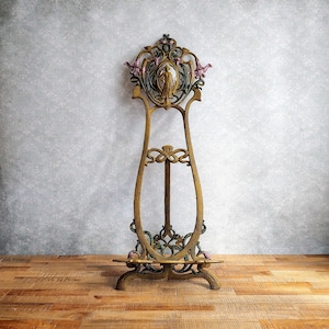 Könnte beinhalten: Ein goldfarbenes Metall-Staffelei mit einem verzierten Design, das eine Frau in einem floralen Motiv zeigt. Die Staffelei hat eine dekorative Oberseite und einen Sockel mit drei Beinen.