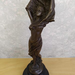 May include: Bronze sculpture of a woman with flowing fabric held above her head. The figure is standing on a black, round base. The sculpture has a dark brown patina and is detailed.