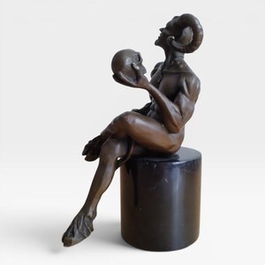 May include: A bronze-toned sculpture of a seated horned figure with cloven hooves, looking upward while holding a human skull in one hand. The figure is positioned on a smooth, cylindrical black base, showcasing detailed musculature and curved horns.