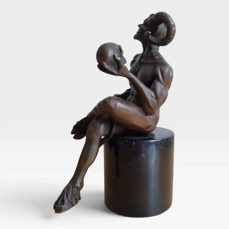 May include: A bronze-toned sculpture of a seated horned figure with cloven hooves, looking upward while holding a human skull in one hand. The figure is positioned on a smooth, cylindrical black base, showcasing detailed musculature and curved horns.