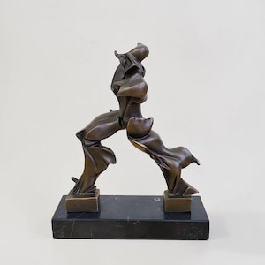 Può includere: Una scultura astratta color bronzo che raffigura una figura dinamica e in movimento con forme fluide e segmentate. La scultura è montata su una base rettangolare in marmo nero, con due piedi ancorati alla superficie.