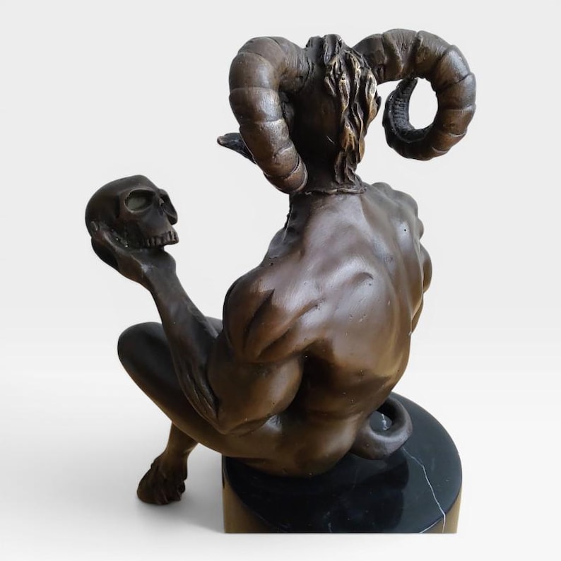 May include: A bronze-toned sculpture of a seated figure with large curved ram horns, muscular back, and a tail, holding a human skull in one hand. The figure is positioned on a cylindrical black marble base with subtle white veining throughout.