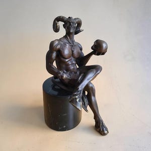 Puede incluir: Una escultura de tono bronce de una figura con cuernos y complexión musculosa, sentada sobre una base cilíndrica de mármol negro. La figura tiene una barba puntiaguda, una cola y pezuñas hendidas, y sostiene un pequeño cráneo redondo y liso.