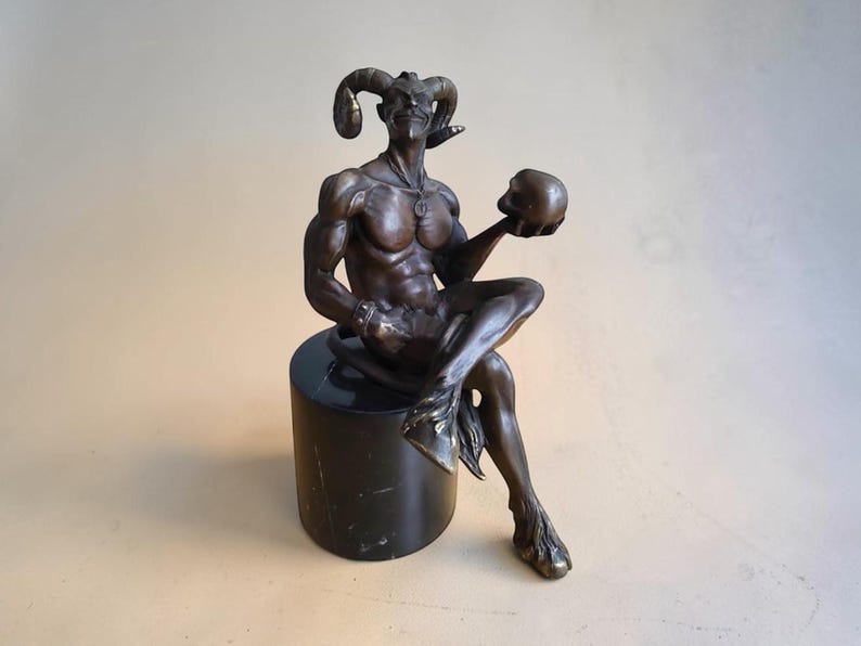 May include: A bronze-toned sculpture of a horned figure with a muscular build, seated on a cylindrical black marble base. The figure has a pointed beard, a tail, and cloven hooves, and is holding a small, smooth, round skull in one hand.