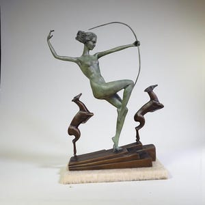 May include: A bronze sculpture depicting a woman with a bow and arrow, in a dynamic pose. Two deer figures are positioned below. The sculpture has a green patina and is mounted on a rectangular base.