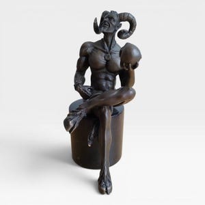 May include: A dark bronze-toned sculpture of a seated horned figure with a muscular physique, cloven hooves, and a tail. The figure holds a spherical object in one hand and wears a pendant necklace, resting upon a cylindrical black base.