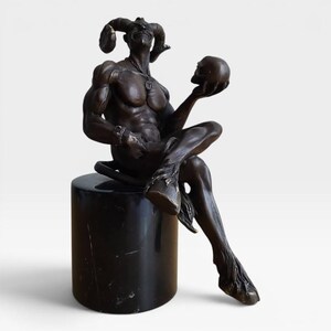 May include: A dark bronze-toned sculpture of a horned, muscular figure with cloven hooves sitting on a cylindrical black marble pedestal. The figure holds a human skull in one hand and wears a necklace with a circular pendant and a wrist cuff.