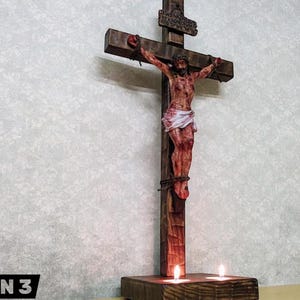 以下が含まれることがあります： イエス・キリストの彫刻が施された木製の十字架。傷と茨の冠が描かれています。十字架は茶色で、人物は白い腰布を着用しています。2本のろうそくが土台に灯っています。「OPTION 3」の文字が下部にあります。
