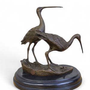 Puede incluir: Escultura de bronce de dos aves con largos picos y patas delgadas, de pie sobre una base en forma de hoja. La escultura está montada sobre una base redonda de mármol negro. Las aves están detalladas con plumas texturizadas.