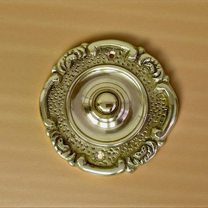Pode incluir: Campainha de latão dourado com um design detalhado de inspiração floral. A campainha tem um botão central e uma borda decorativa. O acabamento em latão confere-lhe um aspeto vintage, adequado para decoração de casa.