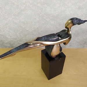 Könnte beinhalten: Eine Bronzeskulptur einer liegenden Figur mit einer länglichen Form. Die Figur wird von einer Kugel getragen, die sich auf einem schwarzen, quadratischen Sockel befindet. Die Skulptur hat eine polierte Metalloberfläche mit Grünspanbereichen.