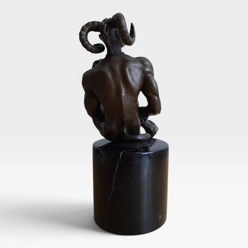 May include: A bronze sculpture of a seated figure with large, curved ram horns, displaying a muscular back and defined anatomy. The figure is mounted on a smooth, cylindrical black marble base with visible white veining throughout the stone.