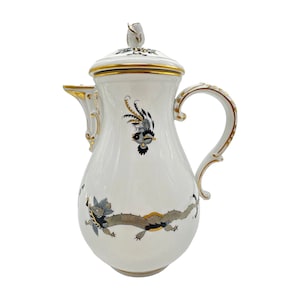 Puede incluir: Cafetera de porcelana blanca con ribete dorado y diseños florales negros y grises. La cafetera tiene una tapa con un pomo dorado y un asa curva.