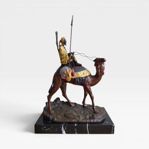 Puede incluir: Escultura de bronce de una persona montada en un camello, sosteniendo una lanza. El jinete viste una túnica amarilla y un turbante. El camello es marrón con mantas de silla decorativas. La escultura está sobre una base de mármol negro.
