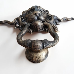 Peut inclure: Un heurtoir de porte en métal couleur bronze foncé représentant une tête de lion sculptée dont la crinière se transforme en deux longs serpents sinueux. Les serpents se courbent vers le bas pour former la poignée fixée à une plaque circulaire.