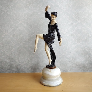 May include: An Art Deco style figurine of a dancing woman in a black dress with a flowing sash, balanced on one leg. The figurine is mounted on a marble base. The sculpture has a cream and black colour scheme.