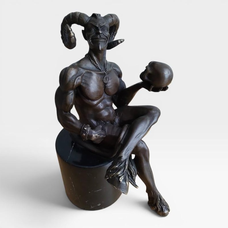 May include: A bronze-toned sculpture of a horned figure with a goatee, sitting on a cylindrical black base. The figure has muscular features, wears a pendant necklace and a wristband, and holds a human skull in one hand while seated.